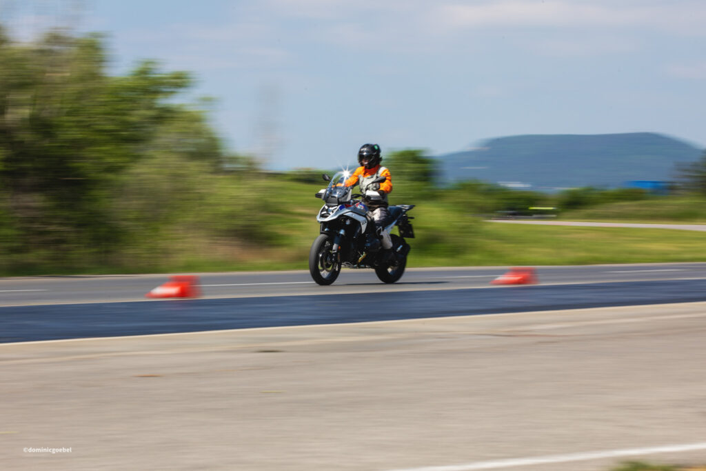 Motorrad Testtage 2026 Teesdorf c Dominic Goebel - I love my moped Motorcyclist in an orange jacket riding a motorcycle on a road, with blurred green trees and hills in the background, conveying speed.