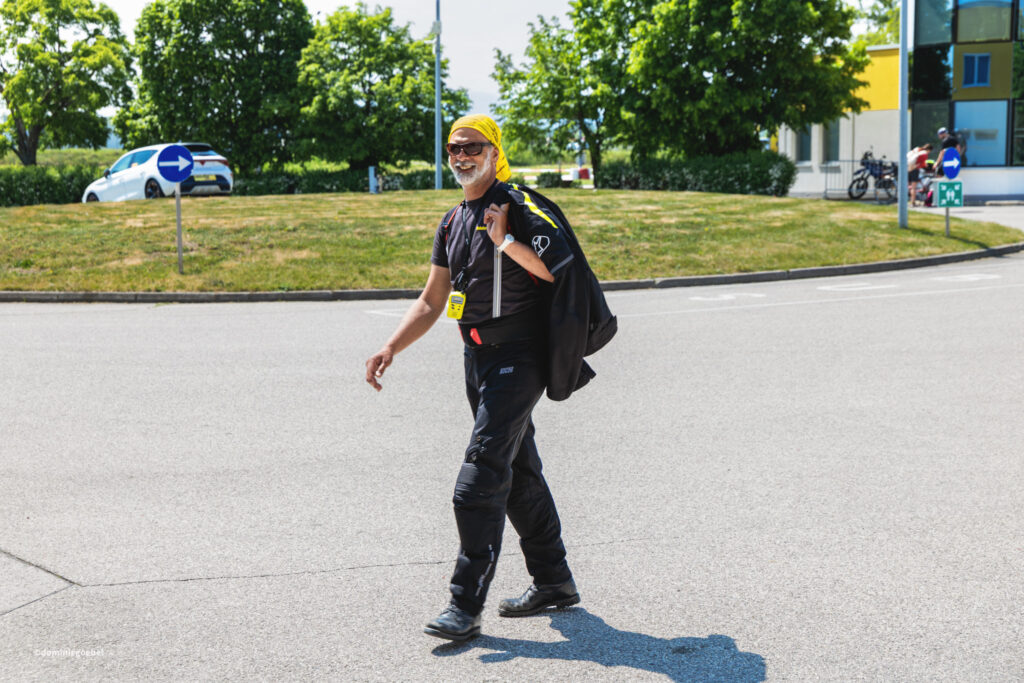 Motorrad Testtage 2026 Teesdorf c Dominic Goebel-11 - I love my moped Man wearing a yellow bandana and black athletic clothes walks across a sunny parking lot, carrying a bag and a yellow device on his chest.