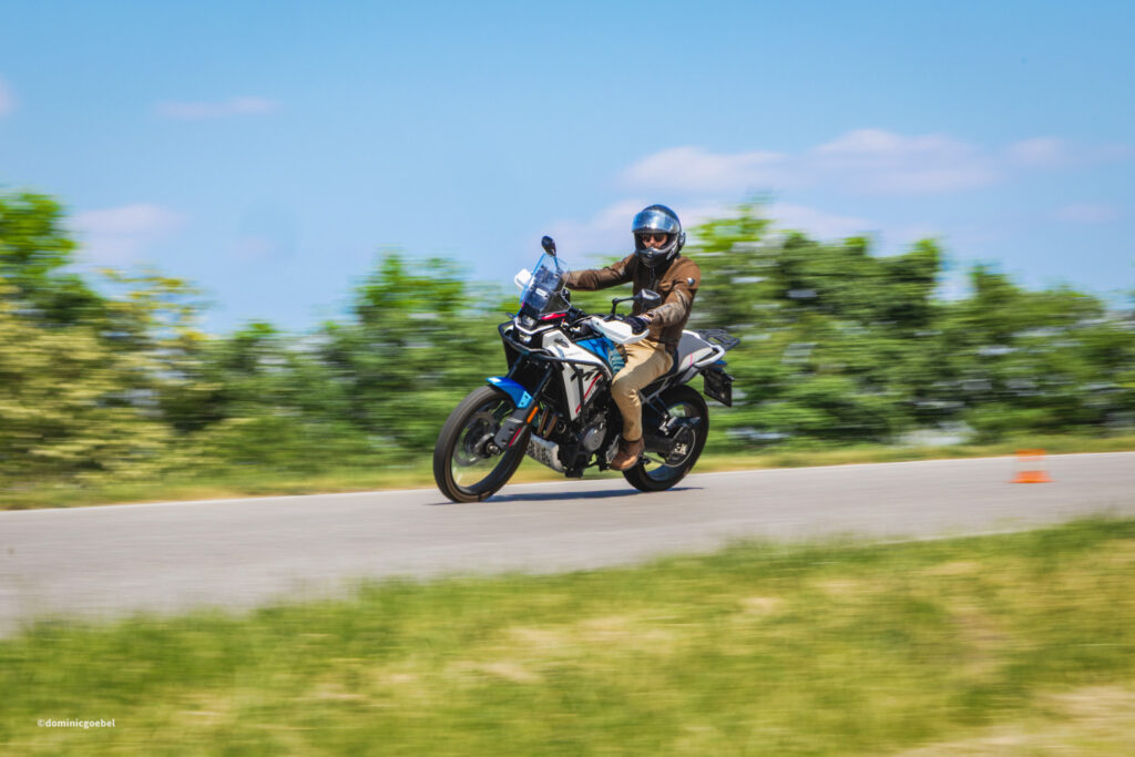 Motorrad Testtage 2026 Teesdorf c Dominic Goebel-14 - I love my moped Motorcyclist wearing a helmet and brown jacket rides a blue-and-white motorcycle on a blurred countryside road.