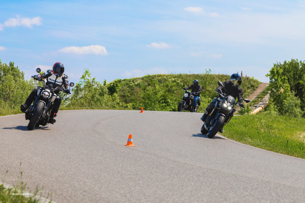 Motorrad Testtage 2026 Teesdorf c Dominic Goebel-15 - I love my moped Three motorcyclists lean into a curve on a rural road, orange cones mark a training course under a blue sky with greenery around.
