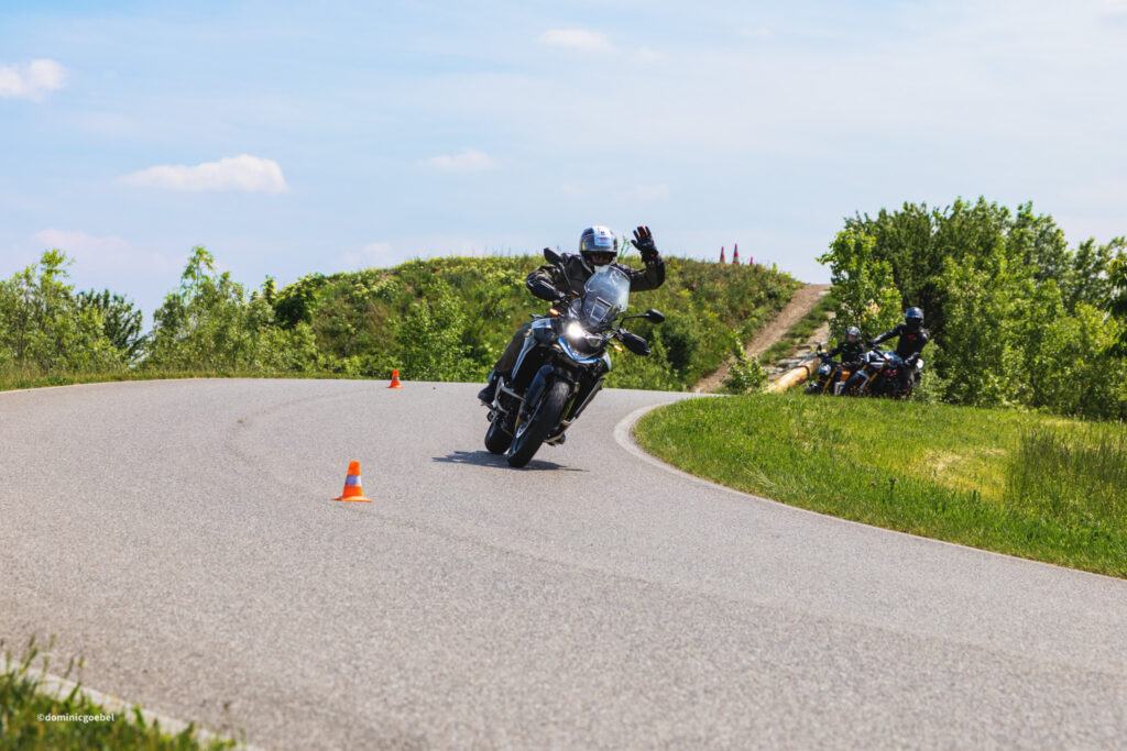 Motorrad Testtage 2026 Teesdorf c Dominic Goebel-16 - I love my moped Motorcyclist leaning into a curve on a sunny rural road, waving as others follow; orange cones mark the course.