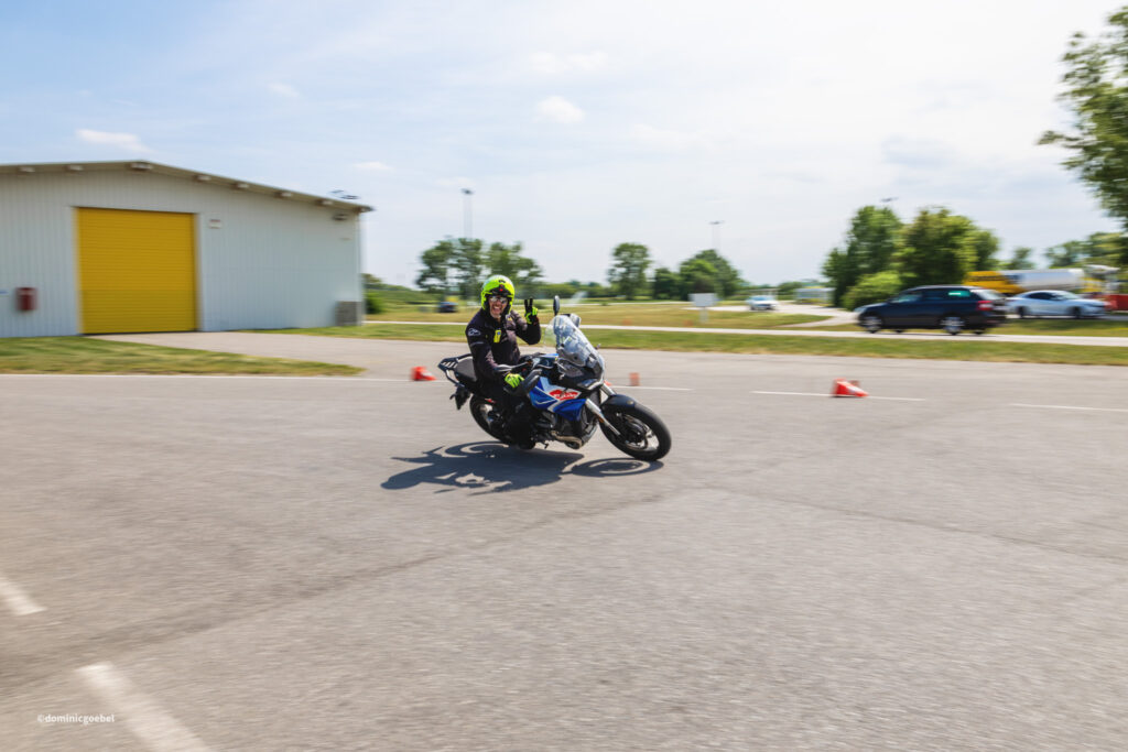Motorrad Testtage 2026 Teesdorf c Dominic Goebel-17 - I love my moped Motorcyclist in neon helmet leaning into a turn on a blue bike, orange cones marking a practice course in a parking lot.