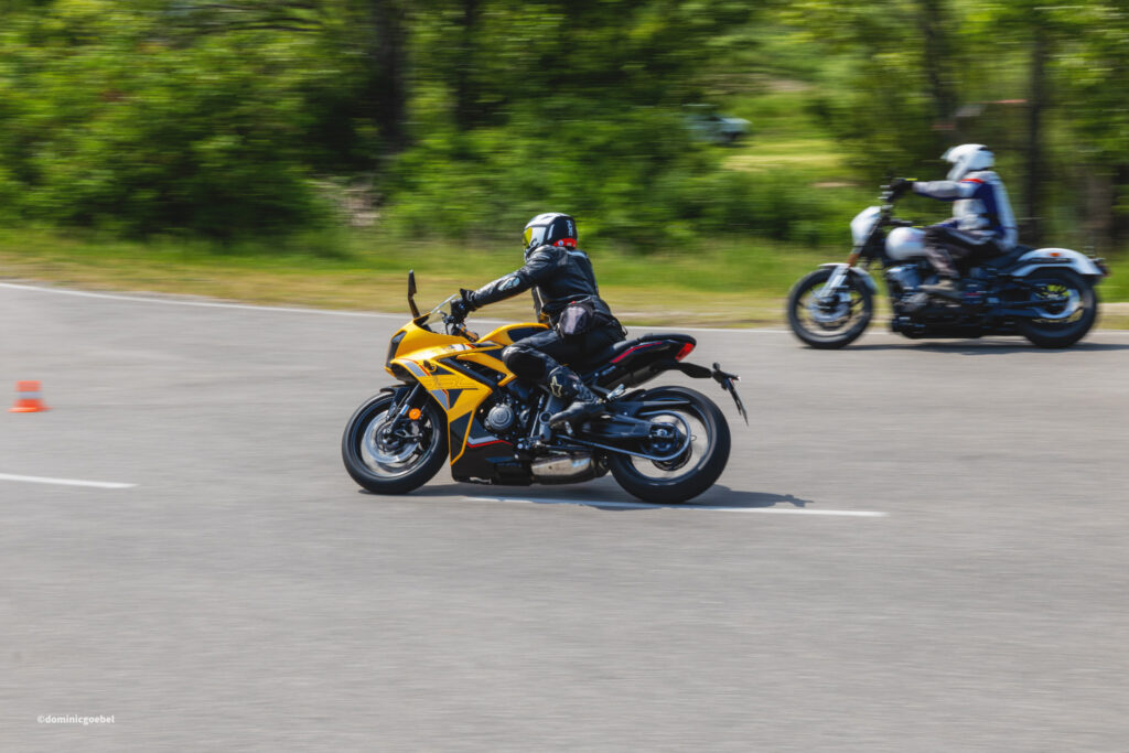 Motorrad Testtage 2026 Teesdorf c Dominic Goebel-19 - I love my moped Motorcyclist on a yellow sport bike leads a two-bike scene on a road, with a white-bike rider in the background and green trees to the rear; orange cones on the left.