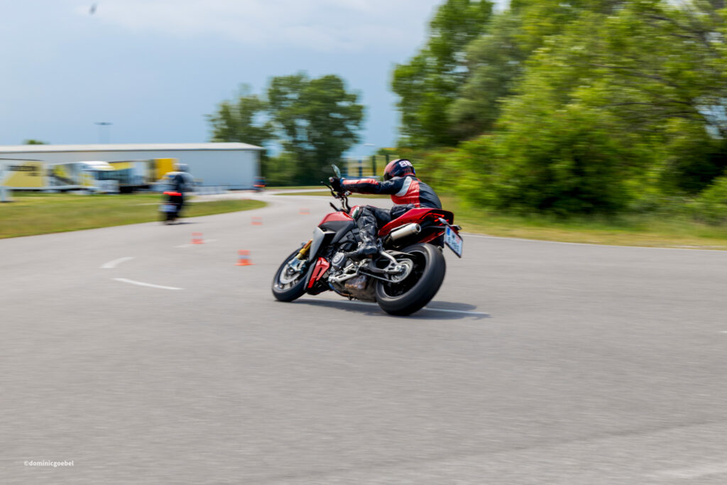 Motorcyclist in red and black gear leaning into a right-hand turn on a sunny road, with blurred trees and buildings in the background.