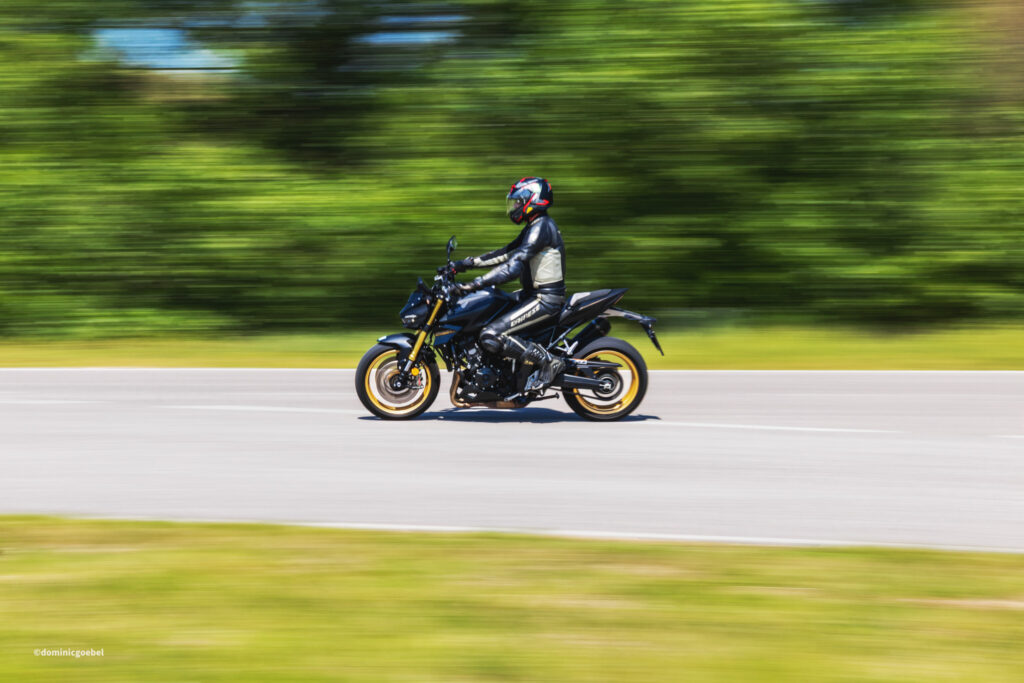 Motorrad Testtage 2026 Teesdorf c Dominic Goebel-31 - I love my moped Motorcycle rider in black protective gear and red helmet speeding along a road, background blurred by motion.