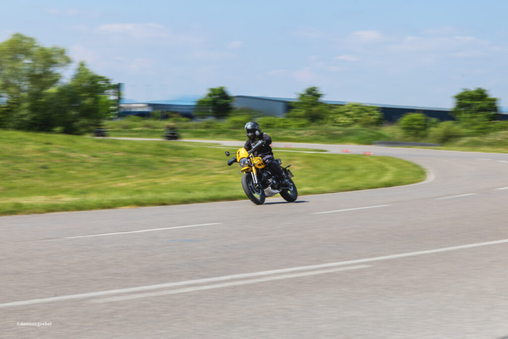 Motorrad Testtage 2026 Teesdorf c Dominic Goebel-34 - I love my moped Motorcyclist on a yellow bike leaning into a curving road, wearing a helmet, in a grassy, industrial background with blue sky.
