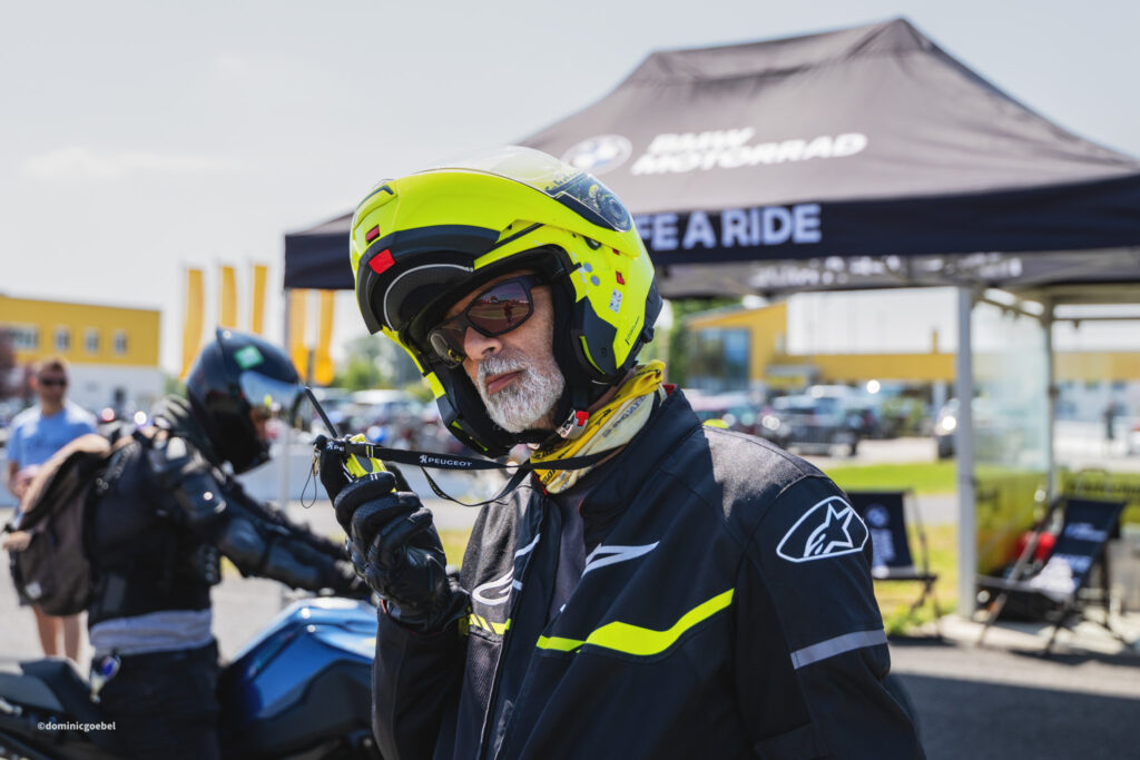 Motorrad Testtage 2026 Teesdorf c Dominic Goebel-36 - I love my moped Close-up of a rider in a bright neon yellow helmet adjusting the strap at an outdoor motorcycle event. Other riders and a tent are in the background.
