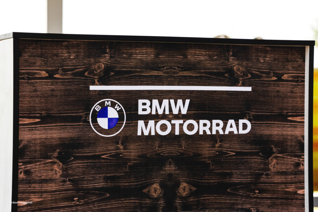 Motorrad Testtage 2026 Teesdorf c Dominic Goebel-37 - I love my moped Signboard featuring the BMW Motorrad logo and the words 'BMW MOTORB...'? Wait, avoid ellipses. Here's a concise set: 1) "Wooden sign with BMW Motorrad logo and bold white text on a dark, weathered plank background." 2) "BMW Motorrad sign on dark stained wood with the circular blue-and-white logo and large white lettering." 3) "Dark wood sign displaying BMW Motorrad branding and logo.