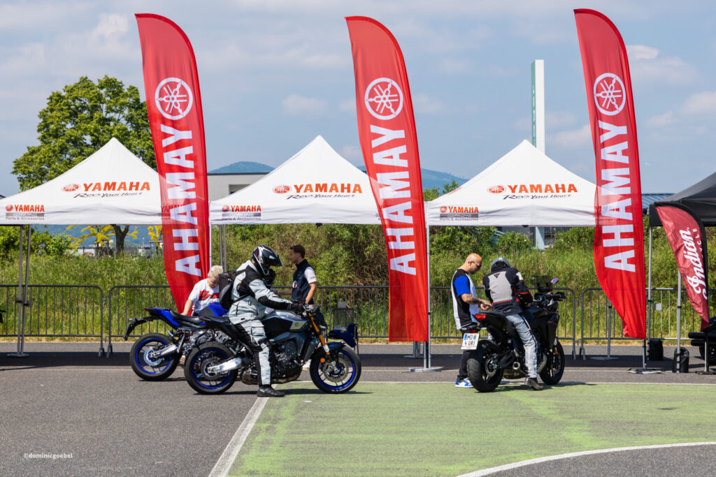 Moto event with riders on blue motorcycles beside white Yamaha tents and tall red flags bearing the Yamaha logo in an outdoor expo setting.