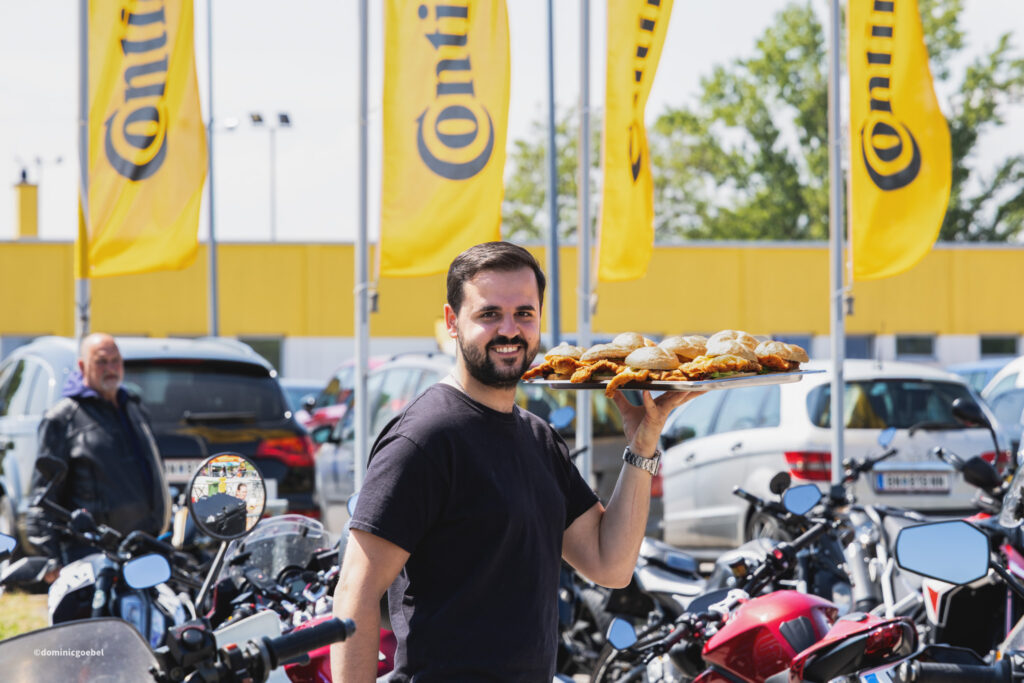 Motorrad Testtage 2026 Teesdorf c Dominic Goebel-41 - I love my moped Man in a black shirt smiles while presenting a tray of pastries outdoors in a parking lot with motorcycles and tall yellow flags in the background.
