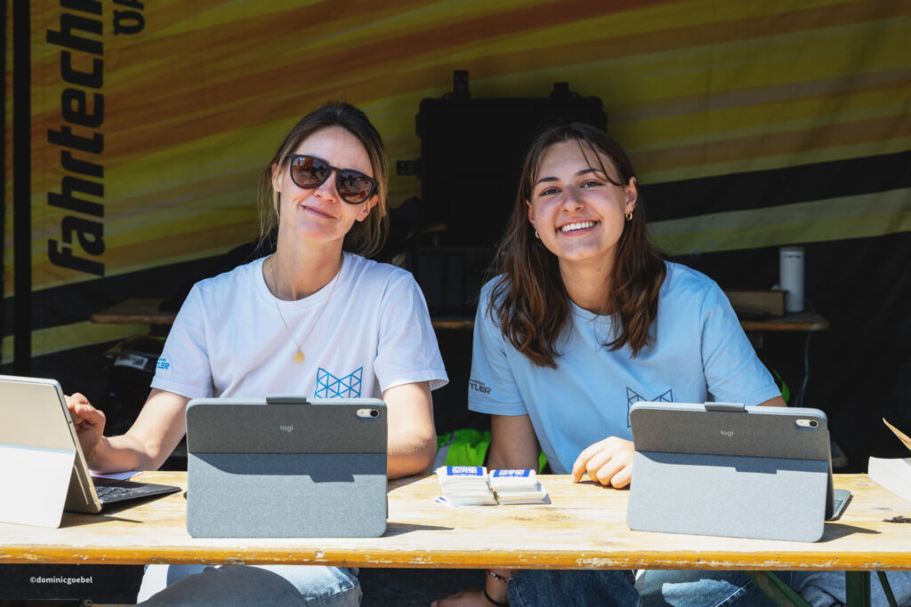 Two women in light blue shirts smile at a booth, with tablets and stacks of business cards on a wooden table.