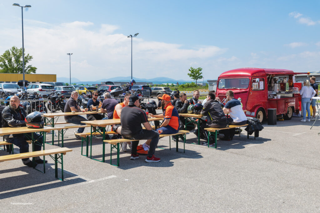 Motorrad Testtage 2026 Teesdorf c Dominic Goebel-8 - I love my moped Motorcycle riders at an outdoor gathering, sitting at wooden tables near a red vintage food truck on a sunny day.
