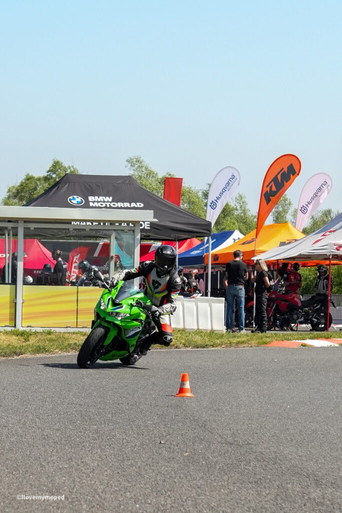 arge2rad-motorradtesttage2026-teesdorf-01 - I love my moped Motorcyclist in protective gear leans into a turn on a bright green sport bike at a motorcycle show, with sponsor tents and an orange cone in the foreground.
