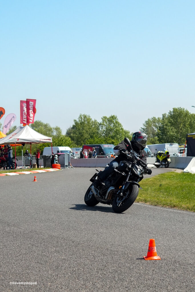 arge2rad-motorradtesttage2026-teesdorf-05 - I love my moped Motorcyclist in black gear leans into a turn on a closed track, orange cones and event tents in the background.