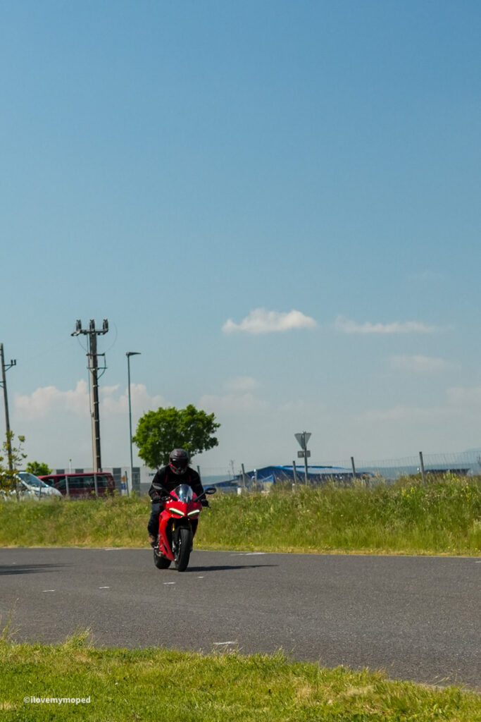 arge2rad-motorradtesttage2026-teesdorf-09 - I love my moped Motorcyclist in black gear riding a red sport motorcycle on a paved road with a grassy field and blue sky behind.