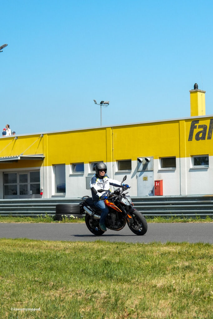 arge2rad-motorradtesttage2026-teesdorf-21 - I love my moped Motorcyclist wearing helmet rides an orange bike on a track with a bright yellow building in the background under a blue sky.