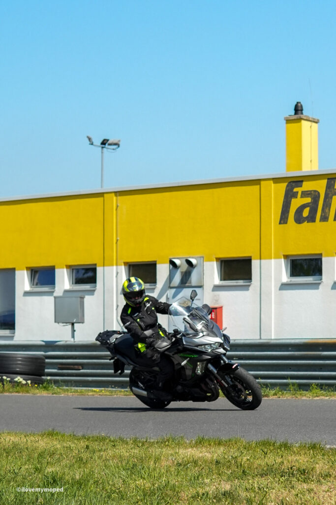 arge2rad-motorradtesttage2026-teesdorf-22 - I love my moped Motorcyclist in black protective gear leaning into a turn on a track, with a bright yellow building in the background.