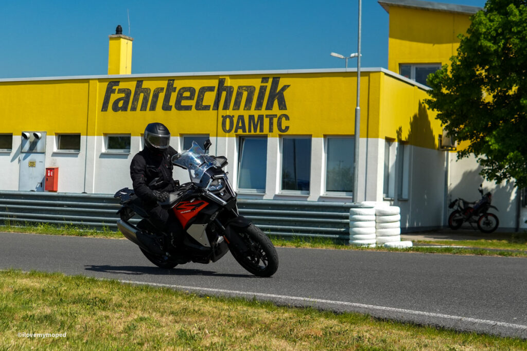 arge2rad-motorradtesttage2026-teesdorf-29 - I love my moped Rider in black gear on a red-and-black motorcycle leaning into a turn on a racetrack with a yellow building behind.