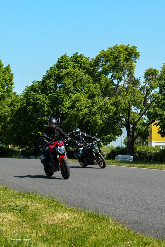 arge2rad-motorradtesttage2026-teesdorf-31 - I love my moped Two motorcyclists leaned into a right-hand turn on a sunny street with green trees in the background.