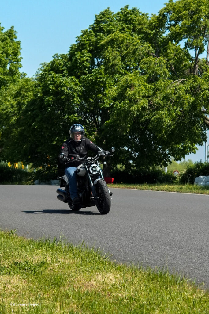 arge2rad-motorradtesttage2026-teesdorf-32 - I love my moped Motorcyclist wearing a helmet and jacket rides a black motorcycle along a curved road with green trees in the background.