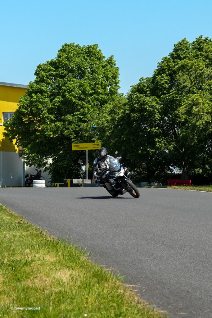 arge2rad-motorradtesttage2026-teesdorf-34 - I love my moped Motorcyclist leaning into a sharp turn on a race track, with green trees and a yellow building behind him.