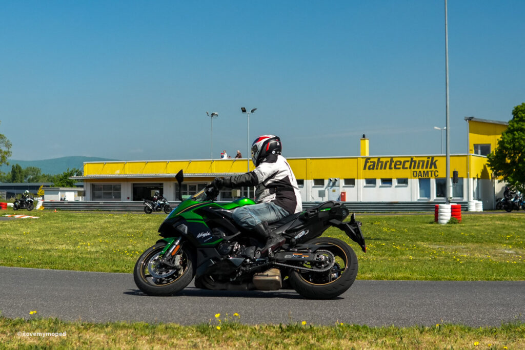 arge2rad-motorradtesttage2026-teesdorf-36 - I love my moped Motorcyclist in white jacket and red helmet riding a green Kawasaki Ninja on a race track with a yellow building in the background, under a blue sky.