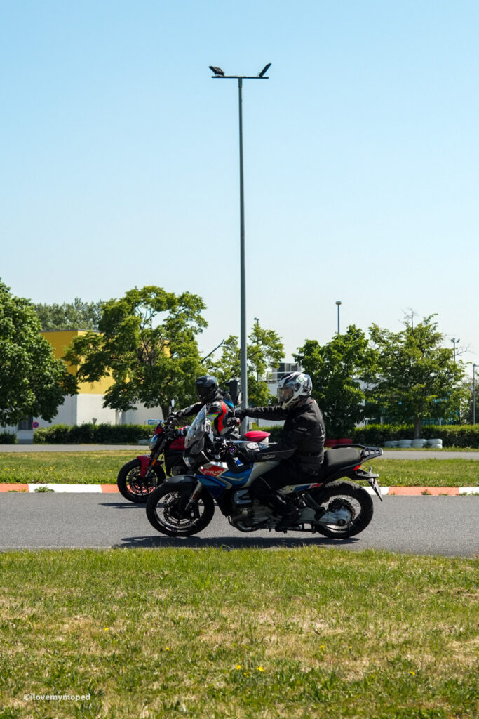 arge2rad-motorradtesttage2026-teesdorf-40 - I love my moped Two motorcyclists wearing protective gear ride on a paved road in a parking lot under a tall streetlight, with trees and a yellow building in the background.