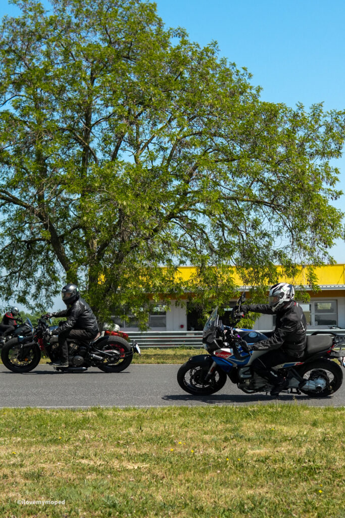 arge2rad-motorradtesttage2026-teesdorf-41 - I love my moped Two motorcyclists ride along a road past a large tree with a yellow building in the background.