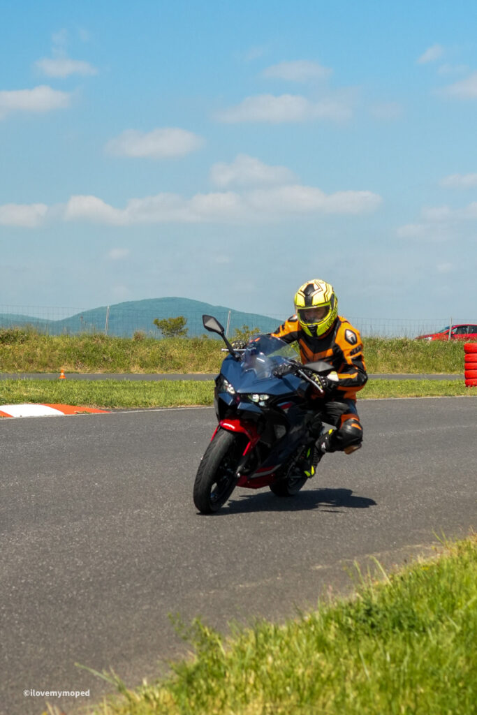 arge2rad-motorradtesttage2026-teesdorf-44 - I love my moped Motorcyclist in orange gear and a yellow helmet leaning into a turn on a racetrack with a blue sky and grassy infield.