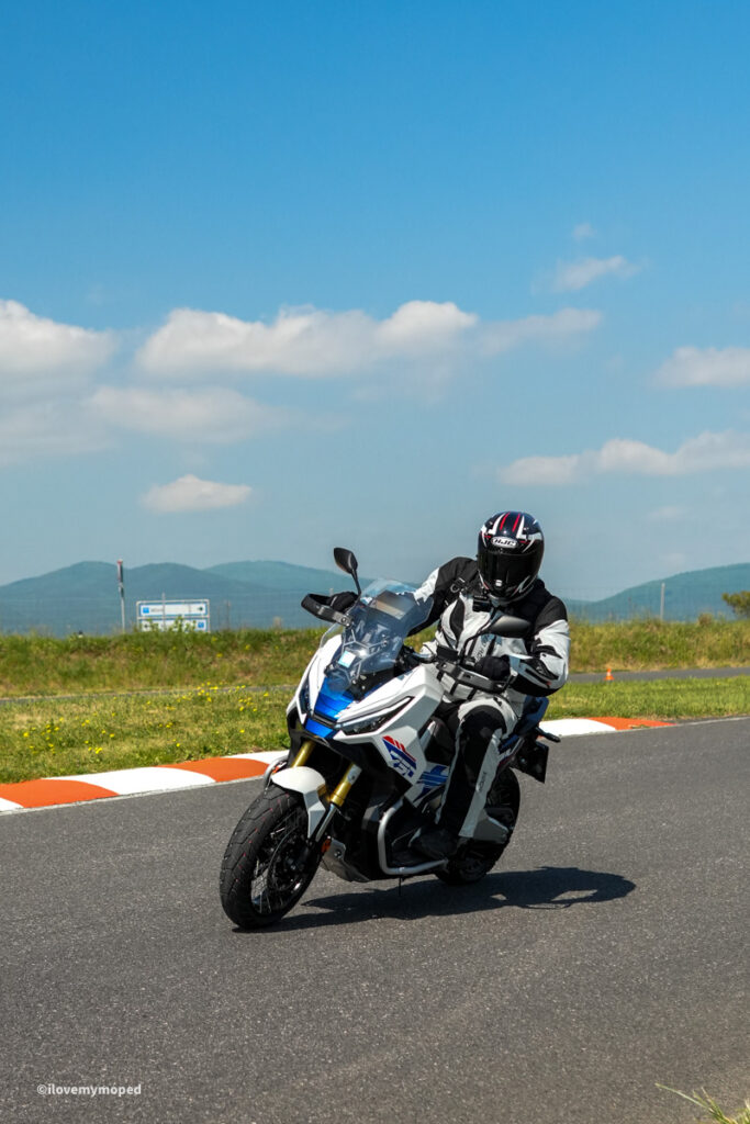 arge2rad-motorradtesttage2026-teesdorf-51 - I love my moped Motorcyclist wearing full gear leans into a turn on a white and blue adventure bike on a paved road with orange-and-white barriers and green fields beyond.
