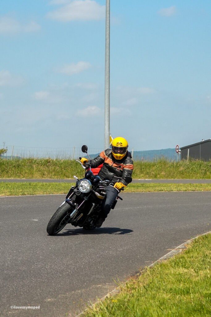 arge2rad-motorradtesttage2026-teesdorf-53 - I love my moped Moto rider in a yellow helmet and black/orange jacket leans into a curve on a dark motorcycle on a paved road, suburban landscape in background.