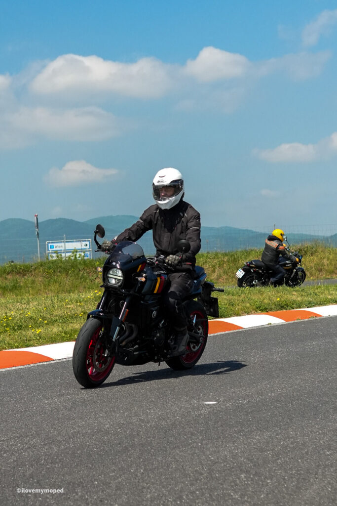 arge2rad-motorradtesttage2026-teesdorf-54 - I love my moped Motorcyclist wearing a white helmet and black riding gear rides a black sport bike on a paved road with an orange-and-white curb; another rider is visible in the distance under a blue sky.