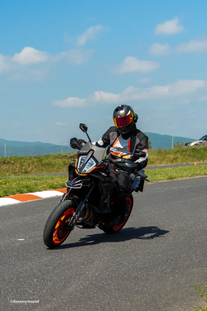 arge2rad-motorradtesttage2026-teesdorf-57 - I love my moped Motorcyclist in black gear and orange helmet rides a sport bike on a curved road with blue sky and grassy fields behind.