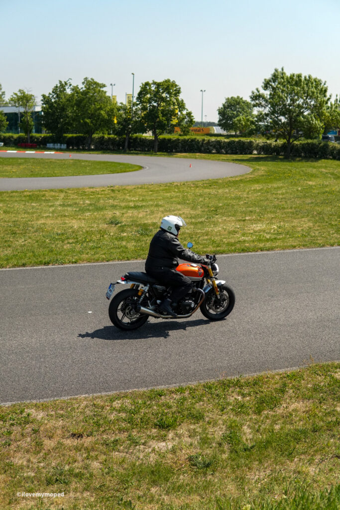 arge2rad-motorradtesttage2026-teesdorf-59 - I love my moped Motorcyclist wearing a white helmet rides an orange motorcycle on a curved paved road with green grass and trees in the background.