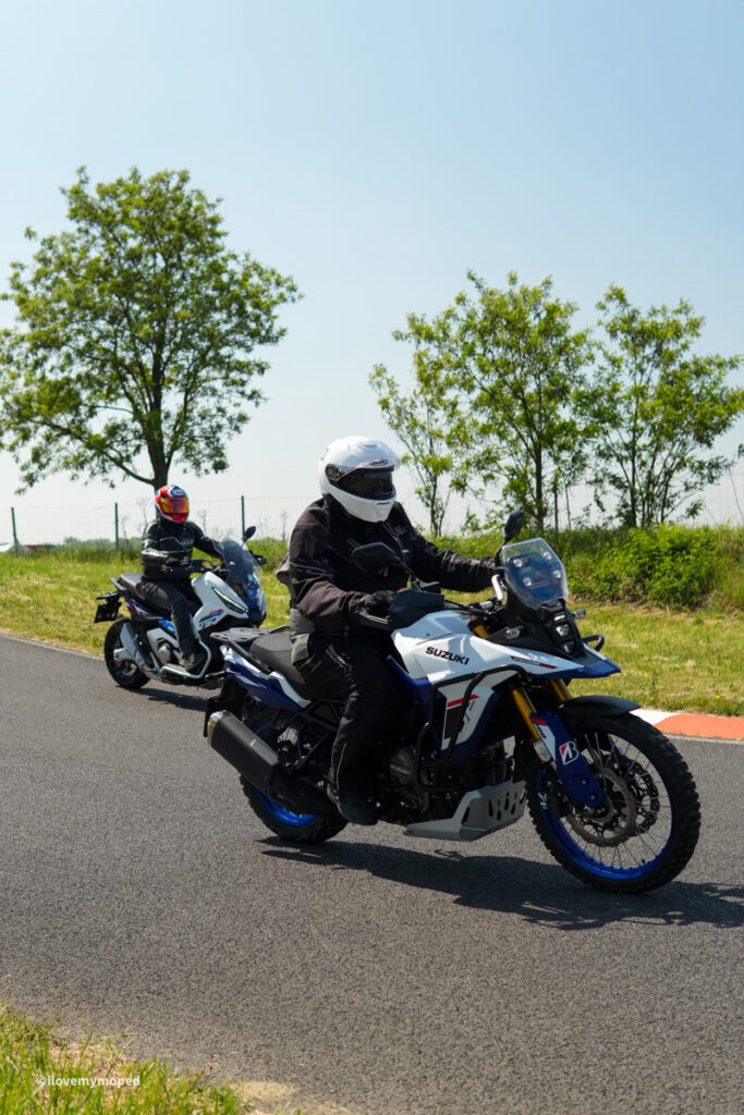 arge2rad-motorradtesttage2026-teesdorf-62 - I love my moped Two motorcyclists ride parallel on a paved road, each wearing full gear and white helmets; the lead rider sits on a white Suzuki adventure bike with blue accents.