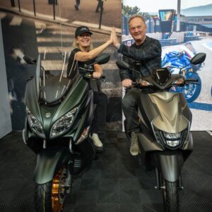 Two people on large scooters in a showroom share a high-five, smiling at the camera with a racing backdrop behind them.