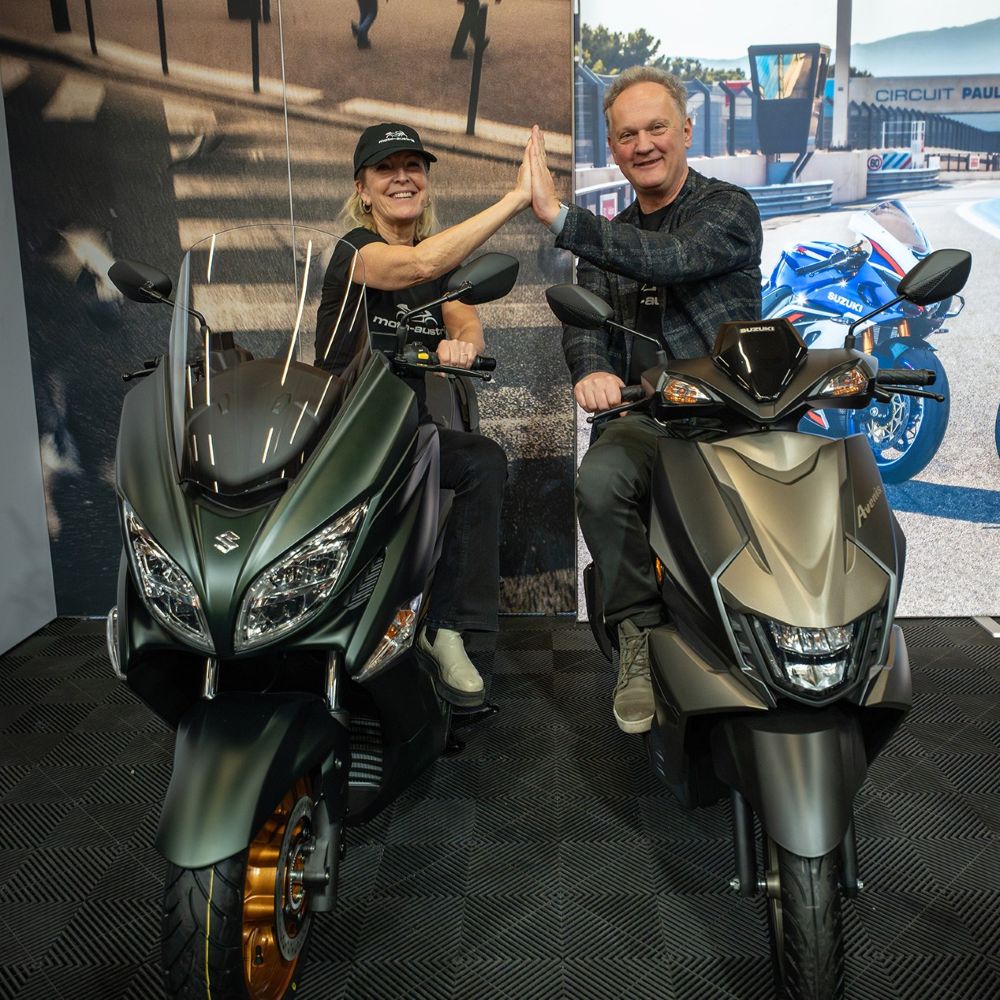 moto-austria 2026 Blogbeitrag I love my Moped - I love my moped Two people on large scooters in a showroom share a high-five, smiling at the camera with a racing backdrop behind them.