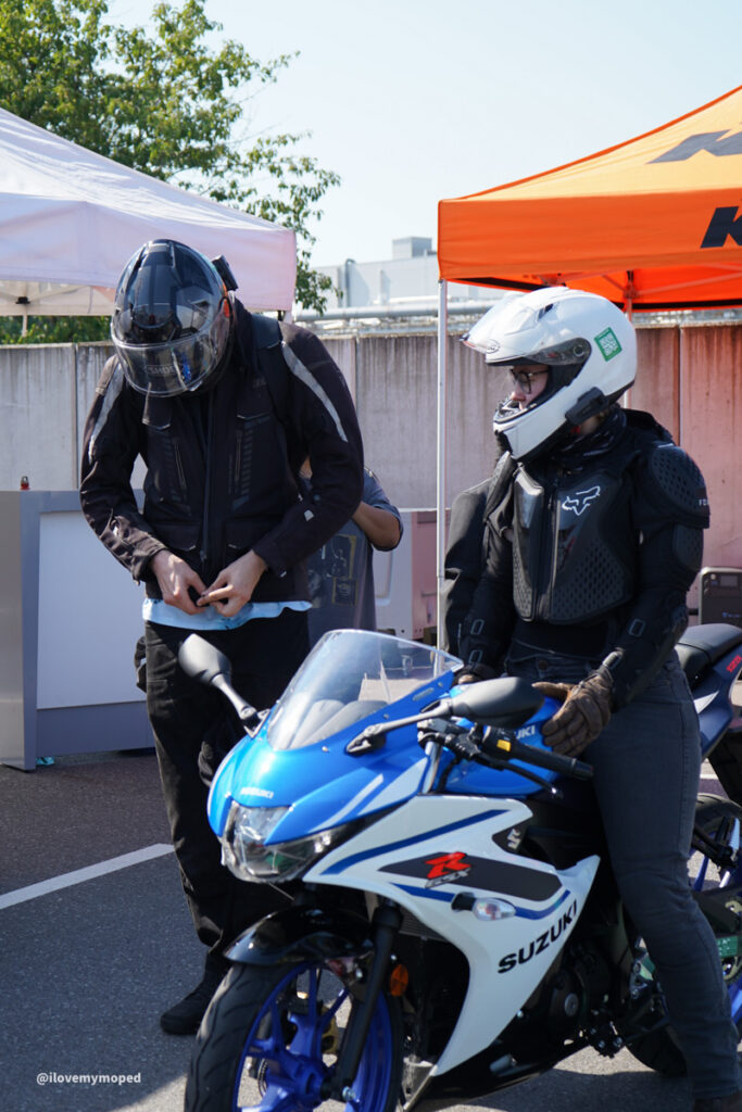 arge2rad-125ertesttag-arböfahrsicherheitszentrumwien-002 - I love my moped Two motorcyclists in gear stand beside a blue Suzuki sport bike under white and orange tents at an outdoor event.