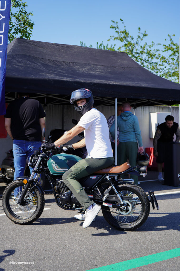 arge2rad-125ertesttag-arböfahrsicherheitszentrumwien-003 - I love my moped Man wearing a white T-shirt and helmet rides a teal vintage motorcycle under a black canopy at an outdoor event.