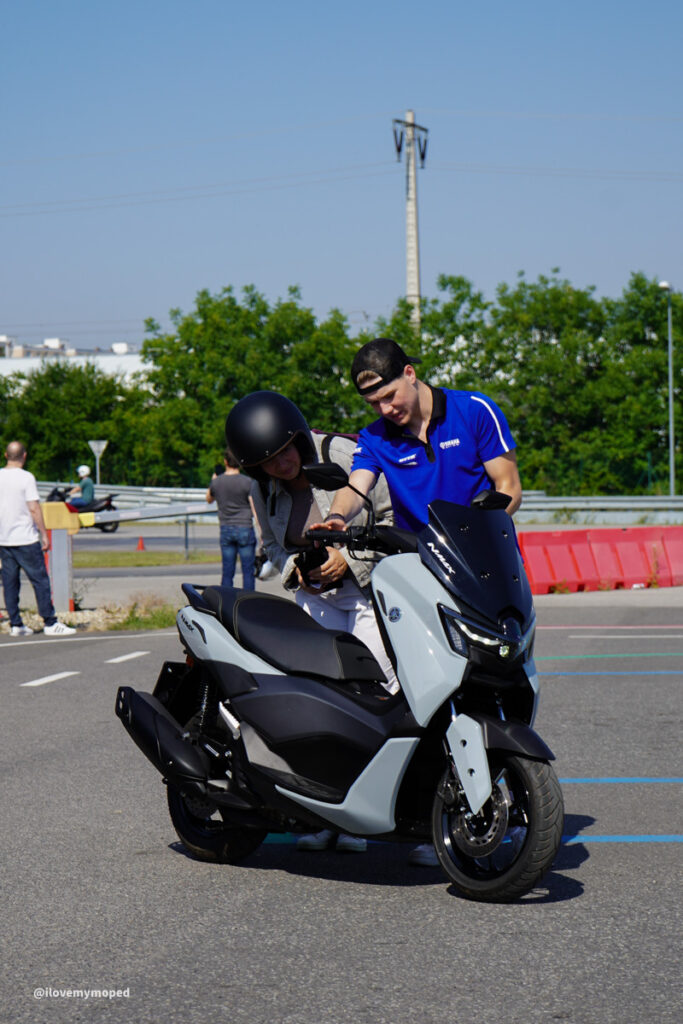 arge2rad-125ertesttag-arböfahrsicherheitszentrumwien-004 - I love my moped Man in a blue shirt helps a woman with a helmet beside a light-blue Yamaha scooter in a parking lot.