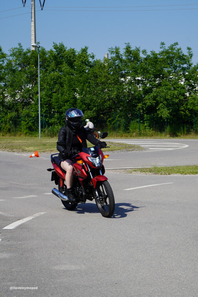 arge2rad-125ertesttag-arböfahrsicherheitszentrumwien-005 - I love my moped Motorcycle rider in black gear and helmet riding a red bike on a paved lot, with a second rider and orange cone in the background.