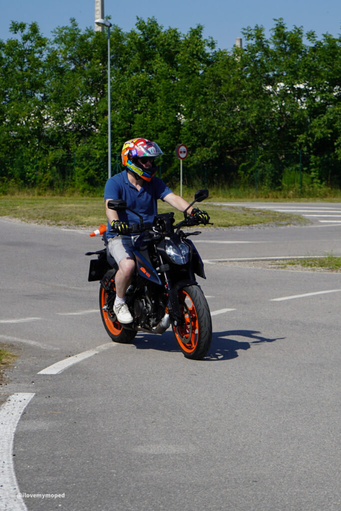 arge2rad-125ertesttag-arböfahrsicherheitszentrumwien-006 - I love my moped Motorcyclist wearing a colorful helmet riding a blue bike with bright orange wheels in a parking lot area.