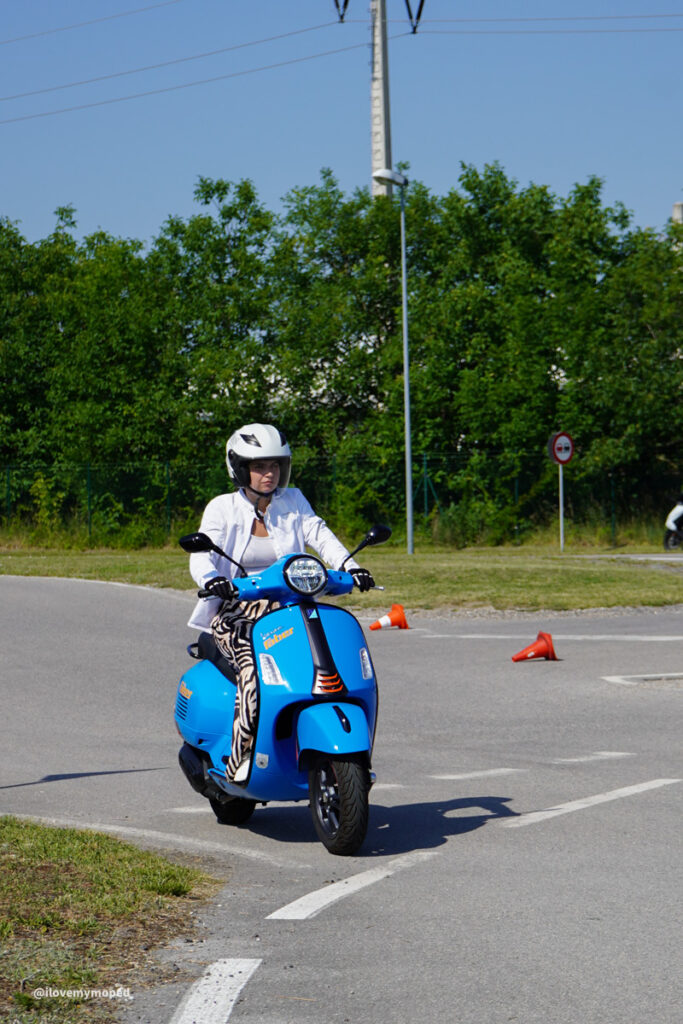 arge2rad-125ertesttag-arböfahrsicherheitszentrumwien-007 - I love my moped Person wearing a white helmet and jacket riding a bright blue scooter on a curved road with orange traffic cones nearby, in a green, sunny area.