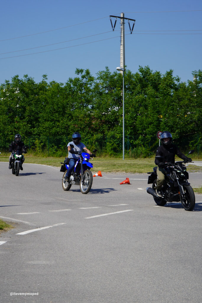 arge2rad-125ertesttag-arböfahrsicherheitszentrumwien-008 - I love my moped Three motorcyclists wearing helmets ride around orange cones on a paved road during a daytime practice or group ride, with a green hedge and power line in the background.