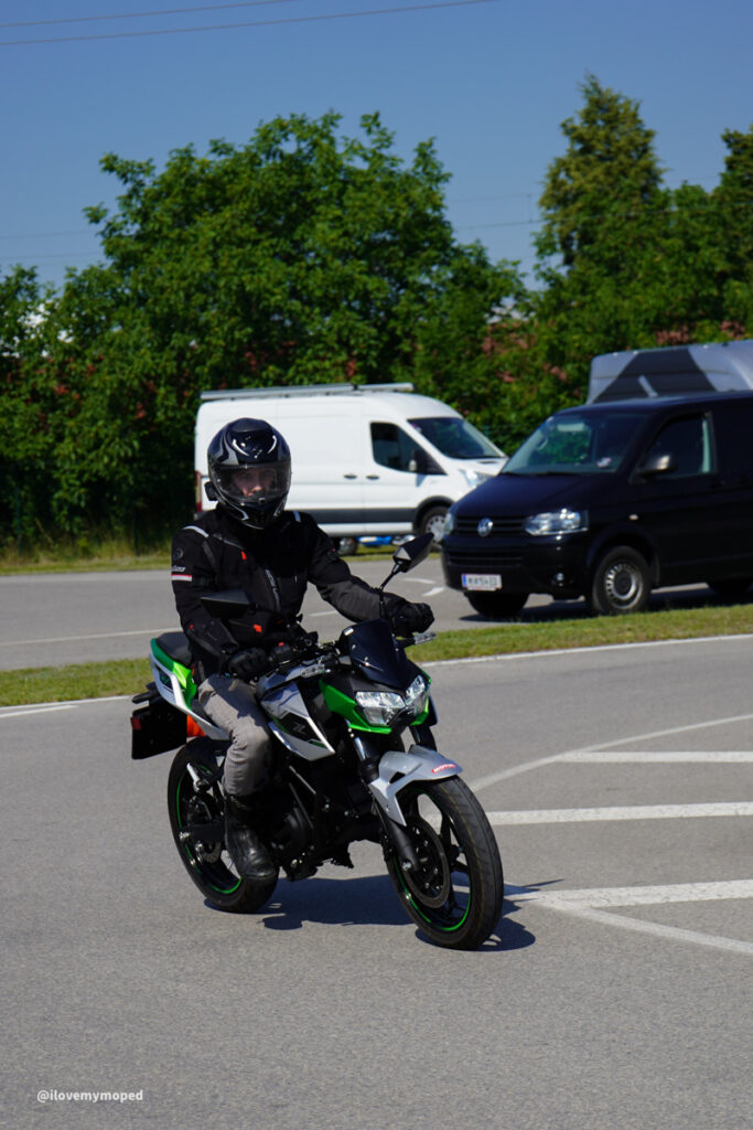 arge2rad-125ertesttag-arböfahrsicherheitszentrumwien-009 - I love my moped Motorcyclist in protective gear riding a green and black motorcycle at an intersection on a sunny day with vans in the background