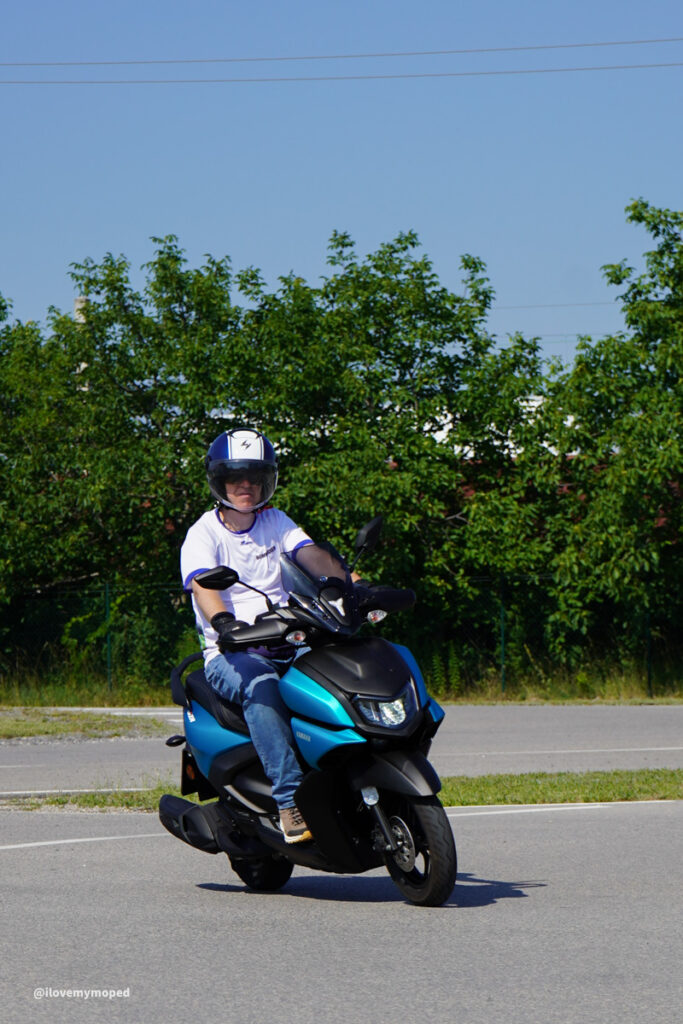 arge2rad-125ertesttag-arböfahrsicherheitszentrumwien-012 - I love my moped Person wearing a helmet rides a blue scooter on a paved road with green trees in the background.
