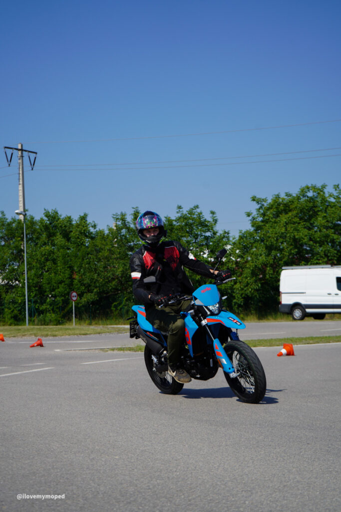 arge2rad-125ertesttag-arböfahrsicherheitszentrumwien-014 - I love my moped Rider in helmet and protective gear on a blue motorcycle navigating an orange-cone course in a paved parking lot.