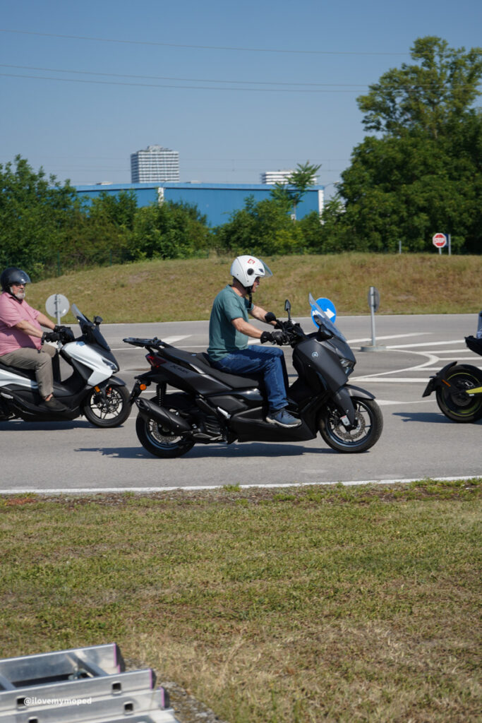 arge2rad-125ertesttag-arböfahrsicherheitszentrumwien-015 - I love my moped Two men ride motor scooters on a sunny road; the front rider wears a teal shirt and white helmet, the rider behind wears pink and a helmet, with a blue industrial building in the background.