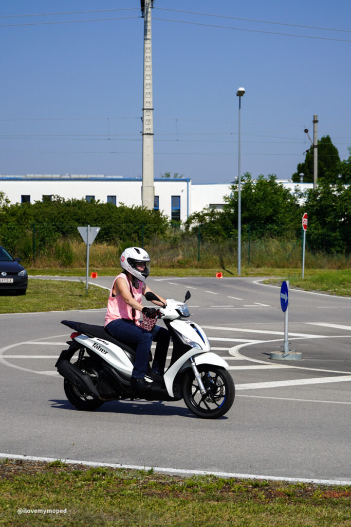 arge2rad-125ertesttag-arböfahrsicherheitszentrumwien-016 - I love my moped Rider on a white scooter wearing a helmet and pink top navigates a roundabout on a sunny day near an industrial area.