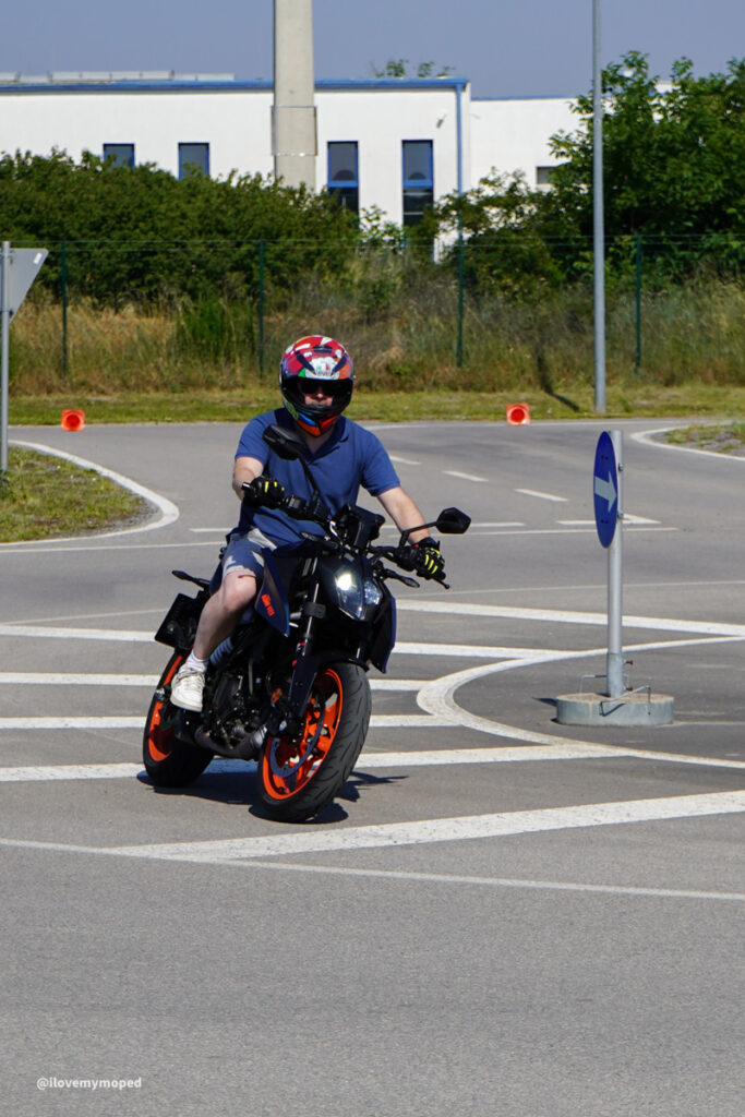 arge2rad-125ertesttag-arböfahrsicherheitszentrumwien-018 - I love my moped Motorcyclist wearing a colorful helmet rides a blue and black motorcycle with orange wheels through a curved road/roundabout area.
